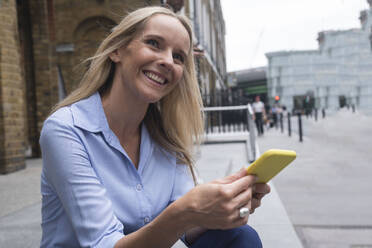 Smiling businesswoman sitting with smart phone in front of building - AMWF01569