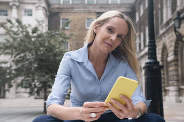 Smiling businesswoman holding smart phone in front of building - AMWF01570