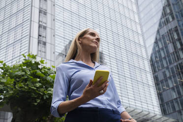 Contemplative businesswoman standing with smart phone in front of office - AMWF01576