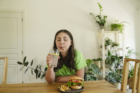 Mädchen trinkt Limonade aus einem Glas mit Burger auf dem Tisch zu Hause - OSF01874