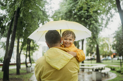 Smiling son with father holding umbrella at park - ANAF01797