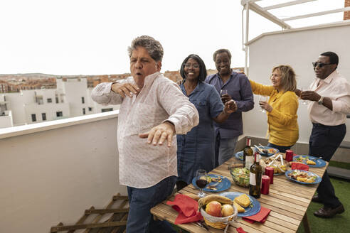 Senior man dancing with friends at dinner party in balcony - JCCMF10657