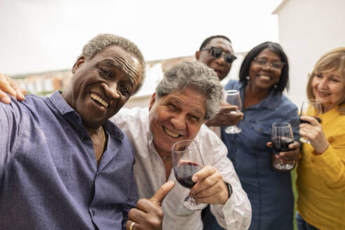 Happy senior man taking selfie with friends enjoying drinks in balcony - JCCMF10667