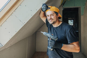 Lächelnder Mann mit Bohrmaschine auf dem Dachboden - VSNF01209