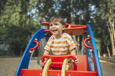 Smiling boy sitting on play equipment in park - ANAF01798