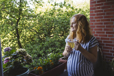 Thoughtful pregnant woman holding herbal tea cup in balcony - IHF01484