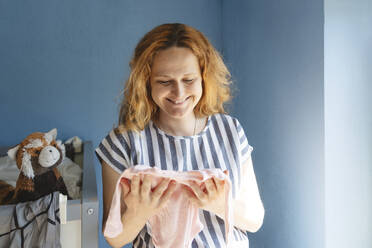 Smiling pregnant woman holding baby clothing at home - IHF01486