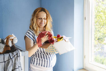Smiling pregnant woman holding box with baby clothing - IHF01488