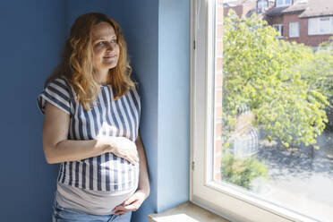 Nachdenkliche schwangere Frau, die zu Hause ihren Bauch berührt - IHF01496