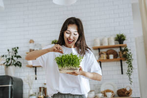 Lächelnde junge Frau mit selbstgezogenen Kräutern in der Küche - NLAF00073