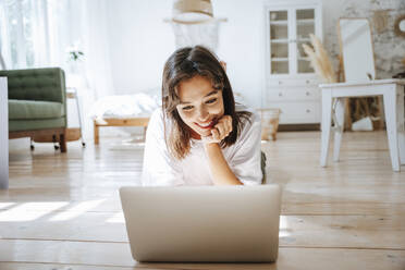 Happy freelancer working with laptop lying on floor at home - NLAF00080