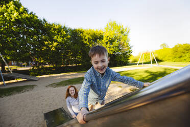 Lächelnder Sohn klettert auf Rutsche mit Mutter im Hintergrund - IHF01509