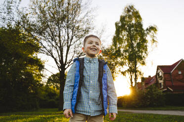 Smiling boy standing in front of house at sunset - IHF01512