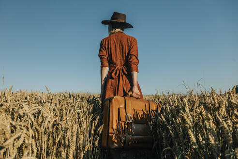 Woman wearing hat holding suitcase in wheat field - VSNF01223