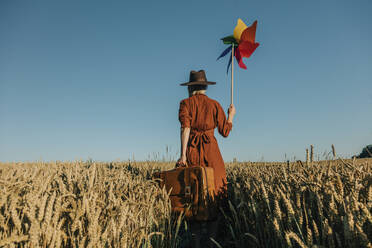 Frau in braunem Kleid hält Windrad-Spielzeug in einem Weizenfeld - VSNF01225