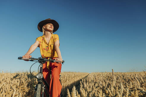 Frau sitzt auf einem Fahrrad in einem Weizenfeld an einem sonnigen Tag - VSNF01232