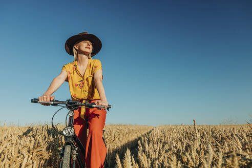 Woman sitting on bicycle in wheat field at sunny day - VSNF01232