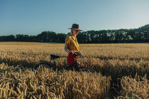 Woman wheeling bicycle in wheat field - VSNF01233