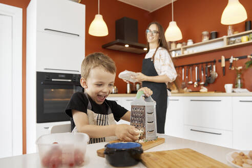 Glücklicher Junge reibt Käse und bereitet mit seiner Mutter zu Hause Essen zu - SANF00131