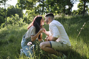 Affectionate couple kissing each other crouching on grass at park - ASGF03897