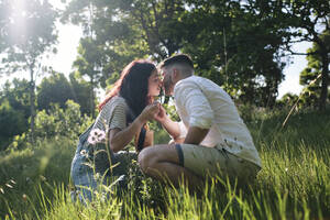 Romantic couple kissing each other crouching on grass at park - ASGF03898