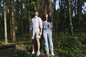 Young couple holding hands standing near tree in forest - ASGF03906