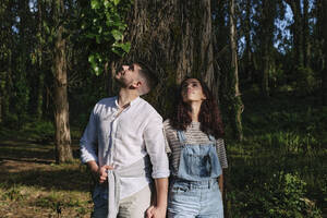 Young couple looking up standing near tree in forest - ASGF03907