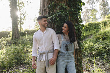 Glückliches Paar, das sich an den Händen hält, vor einem Wald stehend - ASGF03908
