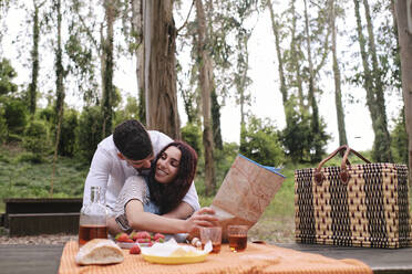 Mann umarmt Frau, die eine Karte hält, die an einem Tisch mit Snacks im Wald sitzt - ASGF03912