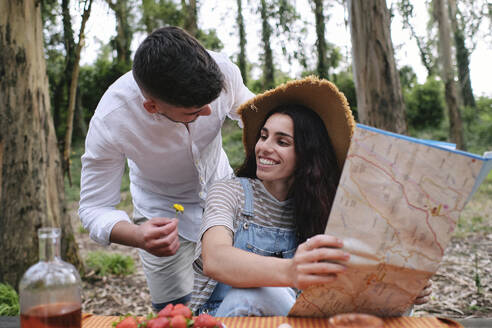 Man giving flower to woman holding map sitting in forest - ASGF03913