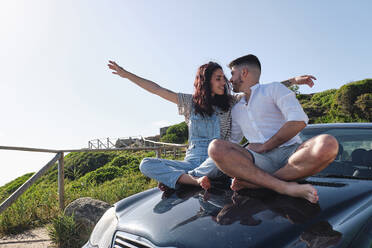 Happy young couple sitting cross-legged on car hood - ASGF03968