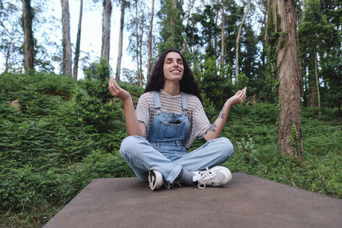 Smiling woman doing Mudra gesture sitting cross-legged in forest - ASGF03992