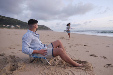 Woman running towards boyfriend sitting on chair at beach - ASGF04006