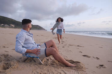 Man sitting on chair looking at girlfriend running on beach - ASGF04007