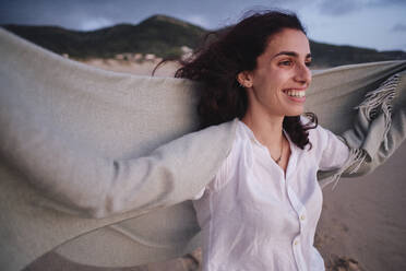Happy woman holding blanket at beach - ASGF04036