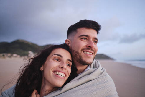 Glückliches junges Paar am Strand in eine Decke gehüllt - ASGF04040