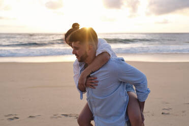 Man giving piggyback ride to girlfriend on beach at sunset - ASGF04053