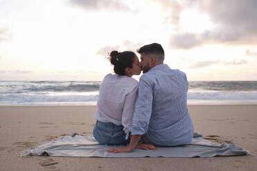 Zärtliches Paar, das sich küsst, während es am Strand im Sand sitzt - ASGF04070