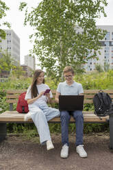 Friends using smart phone and laptop sitting on bench at park - LESF00358