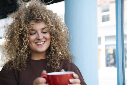 Junge Frau mit lockigem Haar hält Kaffeetasse im Café - AMWF01632