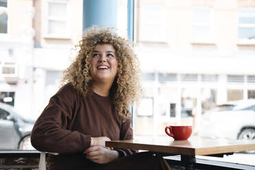 Junge Frau mit lockigem Haar sitzt am Tisch in einem Cafe - AMWF01635