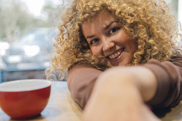 Junge Frau mit lockigem Haar lehnt an einer Kaffeetasse im Café - AMWF01644