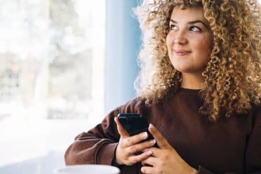 Thoughtful young woman with curly hair holding smart phone at cafe - AMWF01646