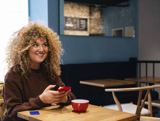 Young woman with curly hair using phone at table in cafe - AMWF01653