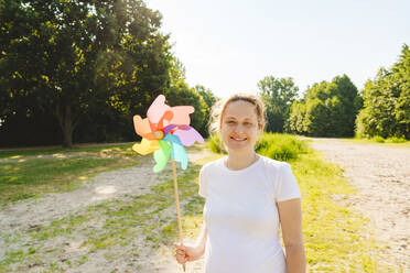 Smiling pregnant woman holding rainbow pinwheel toy on field - IHF01526