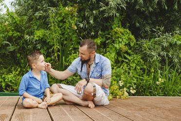 Vater und Sohn sitzen auf einem Steg vor Pflanzen - IHF01541