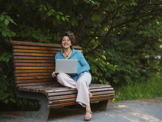 Lächelnde Geschäftsfrau mit Laptop auf einer Parkbank sitzend - IEF00453