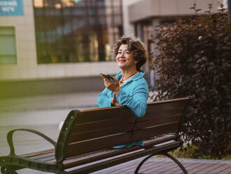 Smiling businesswoman holding smart phone sitting on park bench - IEF00472
