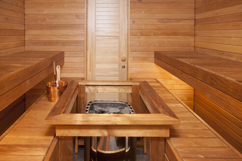 Innenraum eines holzgetäfelten Dampfbads, Sauna, mit zentralem Ofen oder heißem Stein, einem Korb und einer Schöpfkelle für Wasser. - MINF16636
