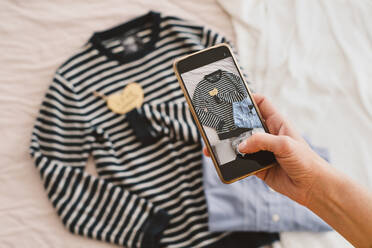 High angle of crop anonymous female taking picture of clothes placed on bed via smartphone in bedroom - ADSF45586